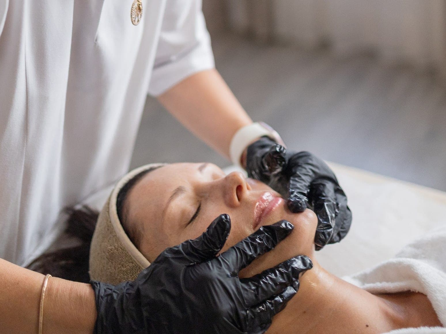 Las manos de la esteticista, con guantes negros, sobre el rostro de una clienta relajada